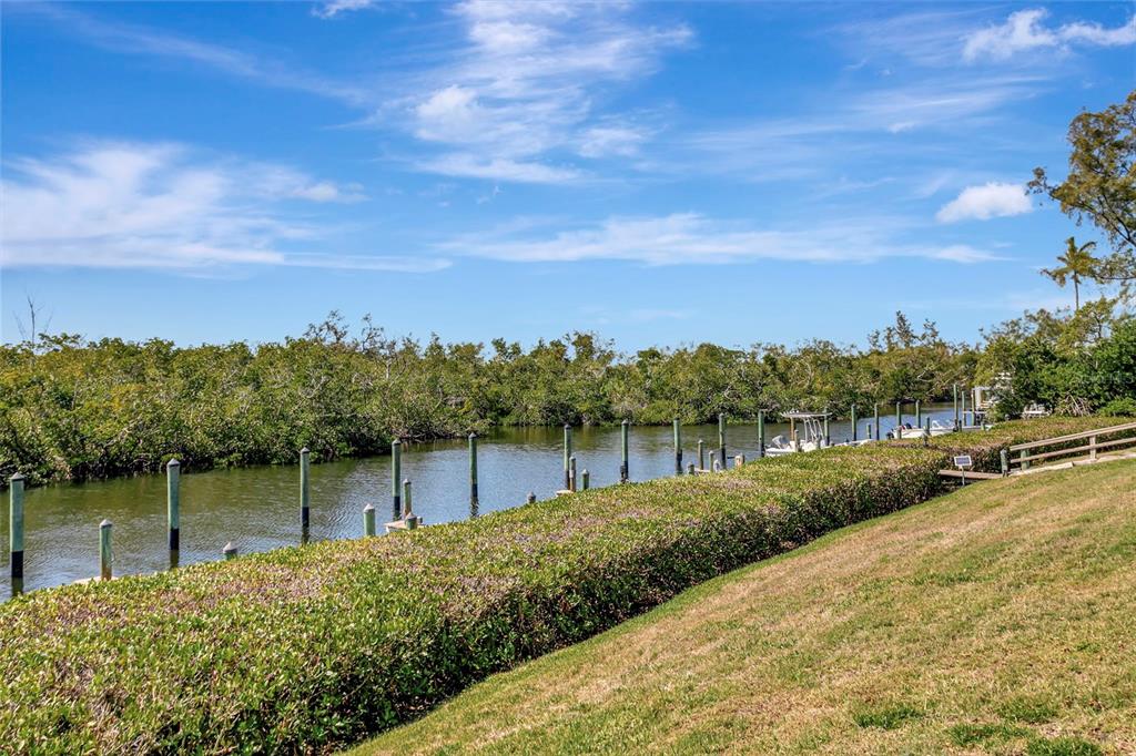 A grassy lawn slopes down toward a canal lined with wooden pilings and dense vegetation — 8734 54TH AVENUE W UNIT 8734, BRADENTON, FL