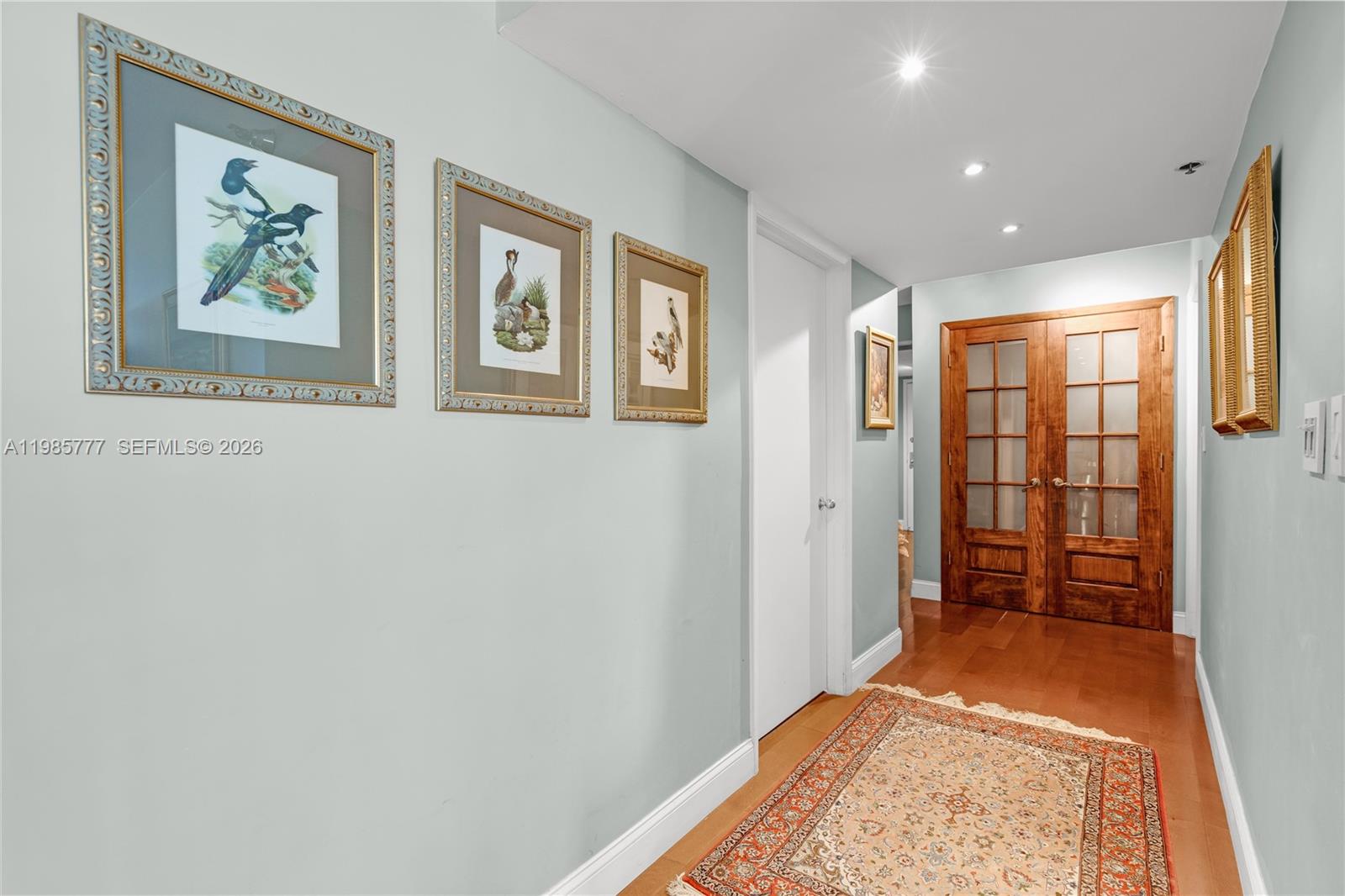 A hallway with light blue walls, framed bird prints, wooden floors, and glass-paneled double doors — 2025 Brickell Ave # 1703, Miami FL 33129, MIAMI, FL