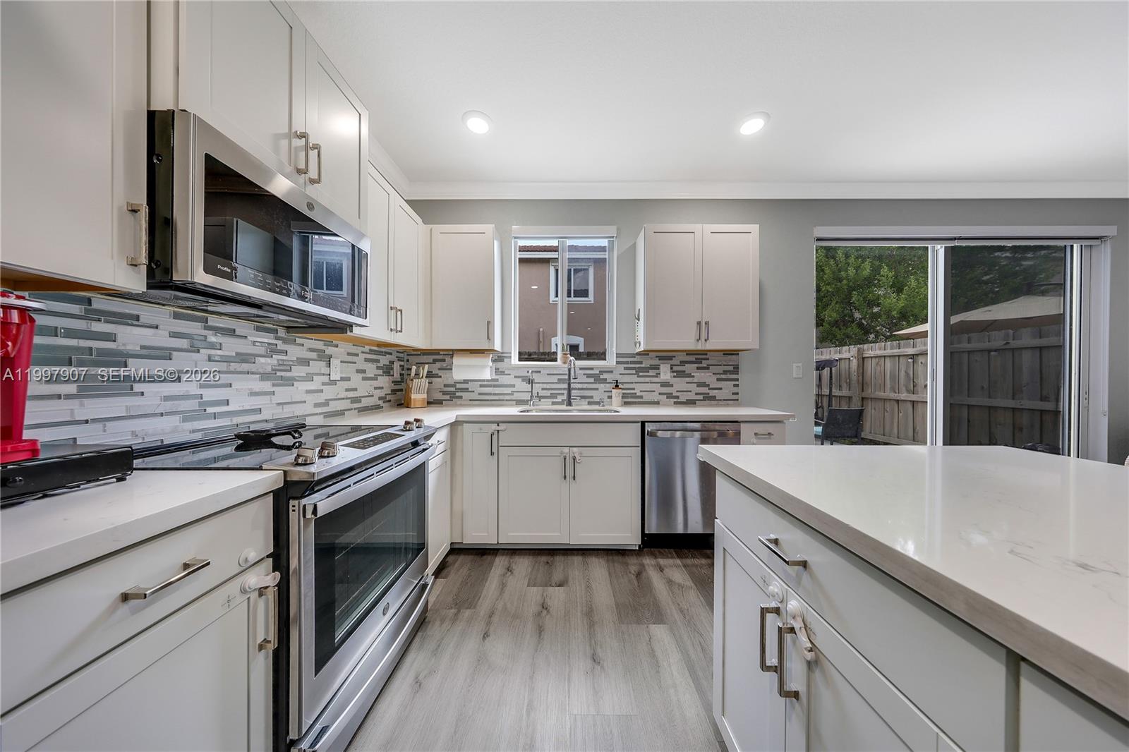 A modern kitchen features white cabinets, stainless steel appliances, a tiled backsplash, and wood flooring — 1079 SE 23rd Ter, Homestead FL 33035, HOMESTEAD, FL