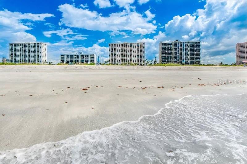A sandy beach with ocean waves in the foreground and tall apartment buildings behind — 1270 GULF BOULEVARD UNIT 304, CLEARWATER, FL