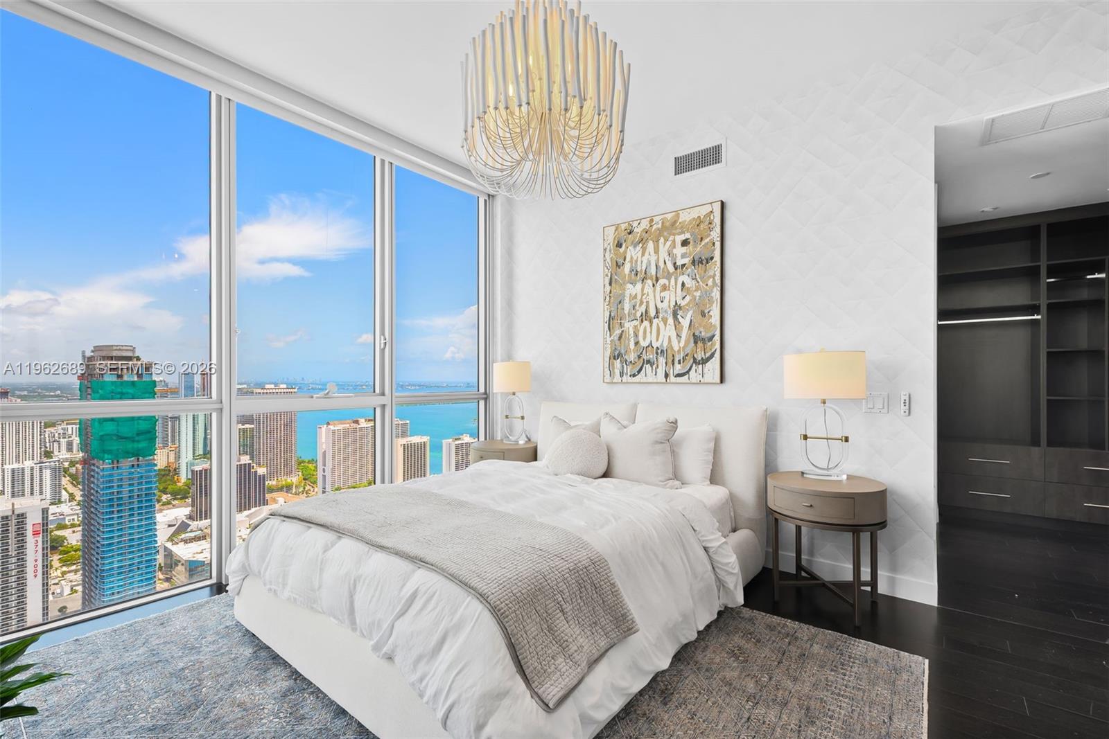 A modern bedroom features a white bed, a large chandelier, and floor-to-ceiling city views — 1100 Biscayne Blvd # 5706, Miami FL 33132, MIAMI, FL