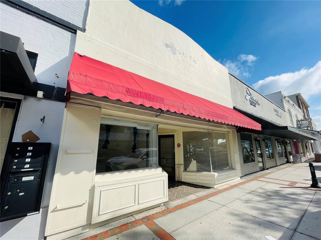 A commercial storefront with a red fabric awning and large display windows on a sidewalk — 109 BROADWAY, KISSIMMEE, FL
