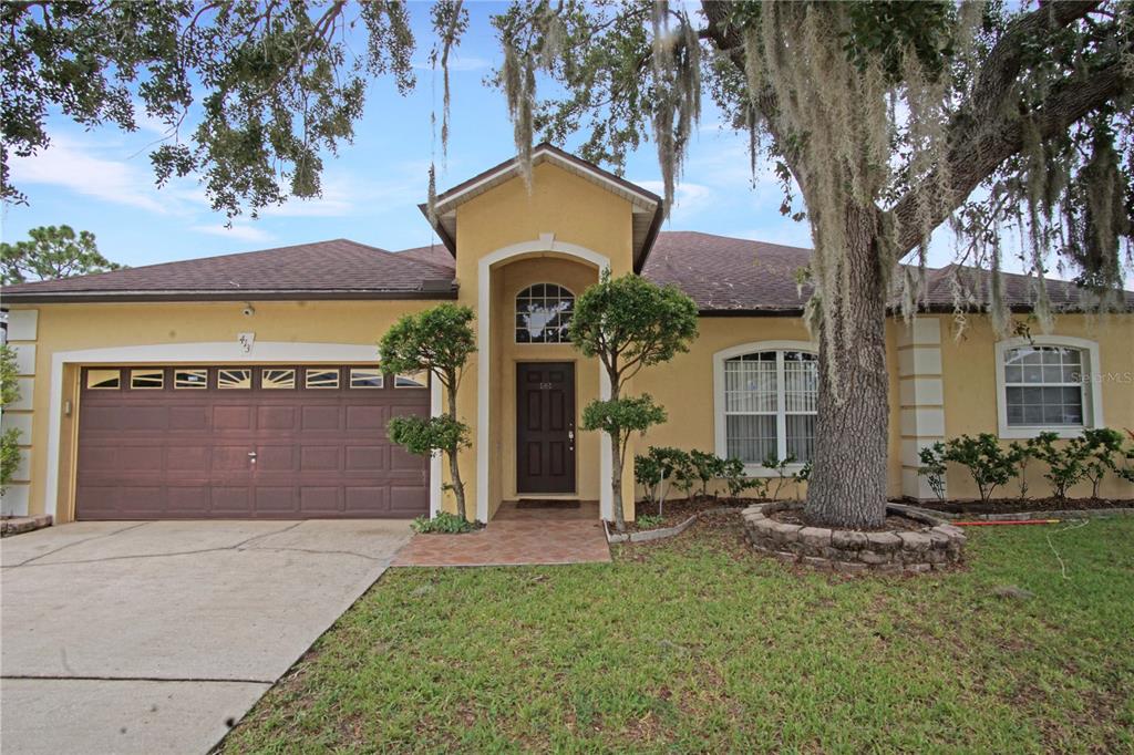 A yellow single-story house with a brown garage door, arched entryway, and a large tree — 413 MARLBERRY LEAF AVENUE, KISSIMMEE, FL
