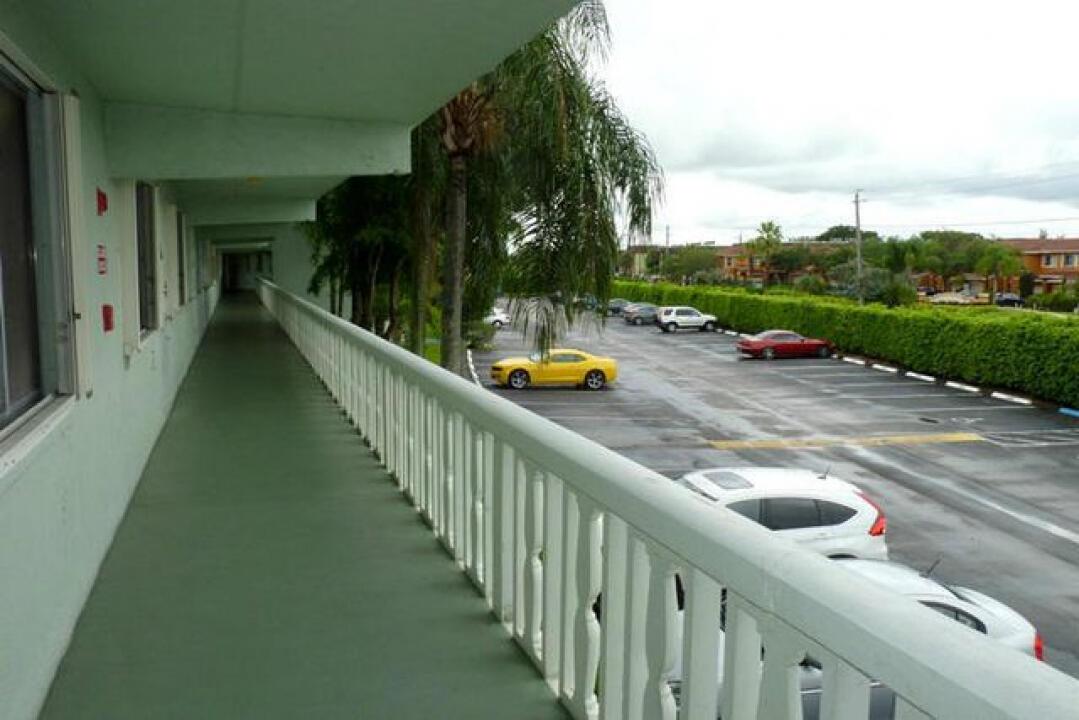 An elevated outdoor walkway overlooks a parking lot with several cars and a hedge — 3300 Springdale Boulevard # 306, Palm Springs FL 33461, PALM SPRINGS, FL