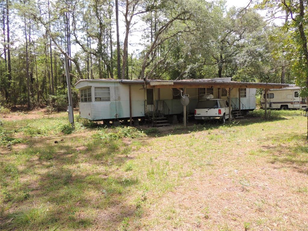 A weathered mobile home with a carport sits on a grassy lot among tall trees — 250 NE 131ST AVENUE, WILLISTON, FL