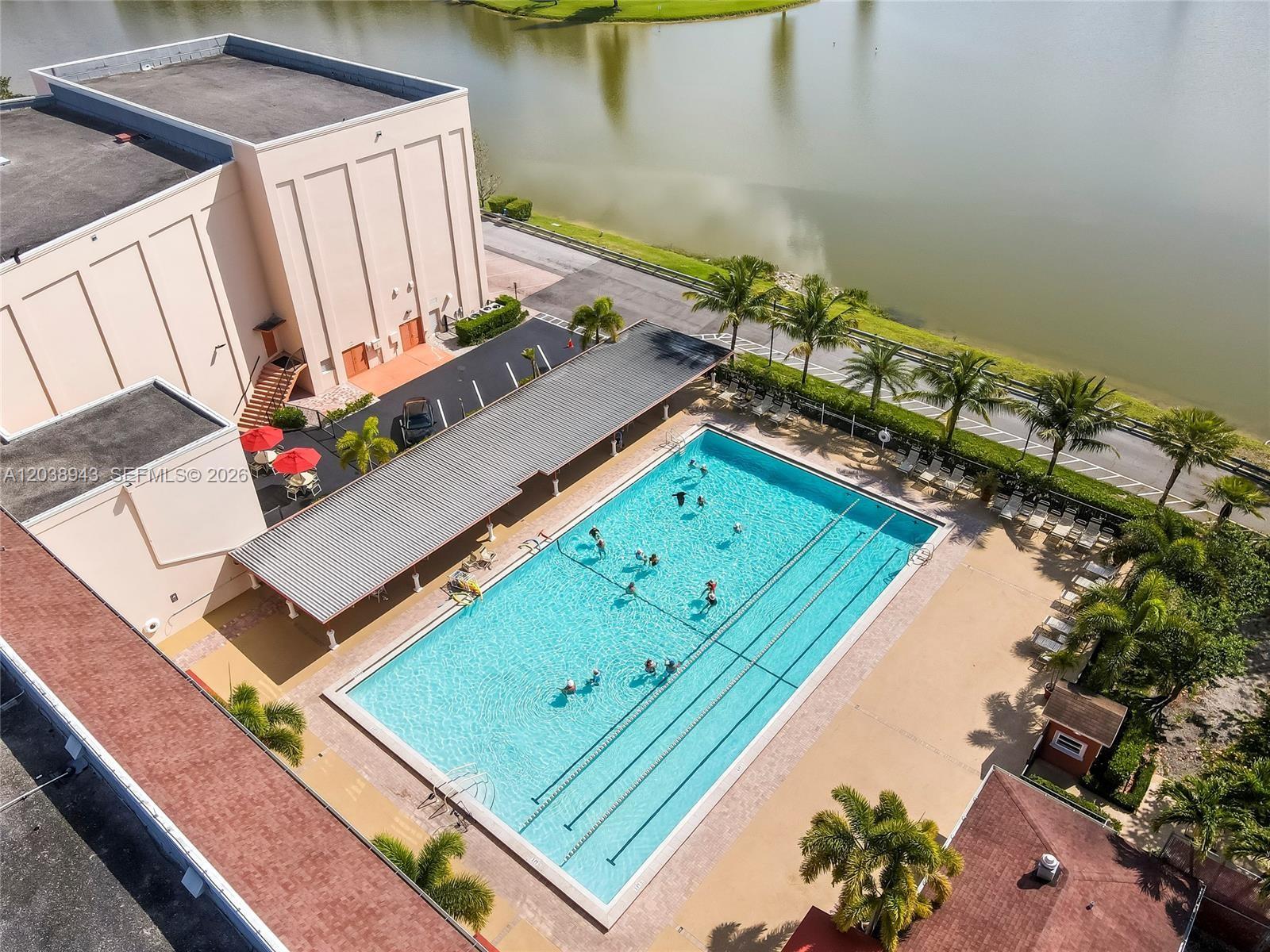 An aerial view shows a rectangular swimming pool with people, a covered patio, and a lake — 82 Dorset B # B, Boca Raton FL 33434, BOCA RATON, FL