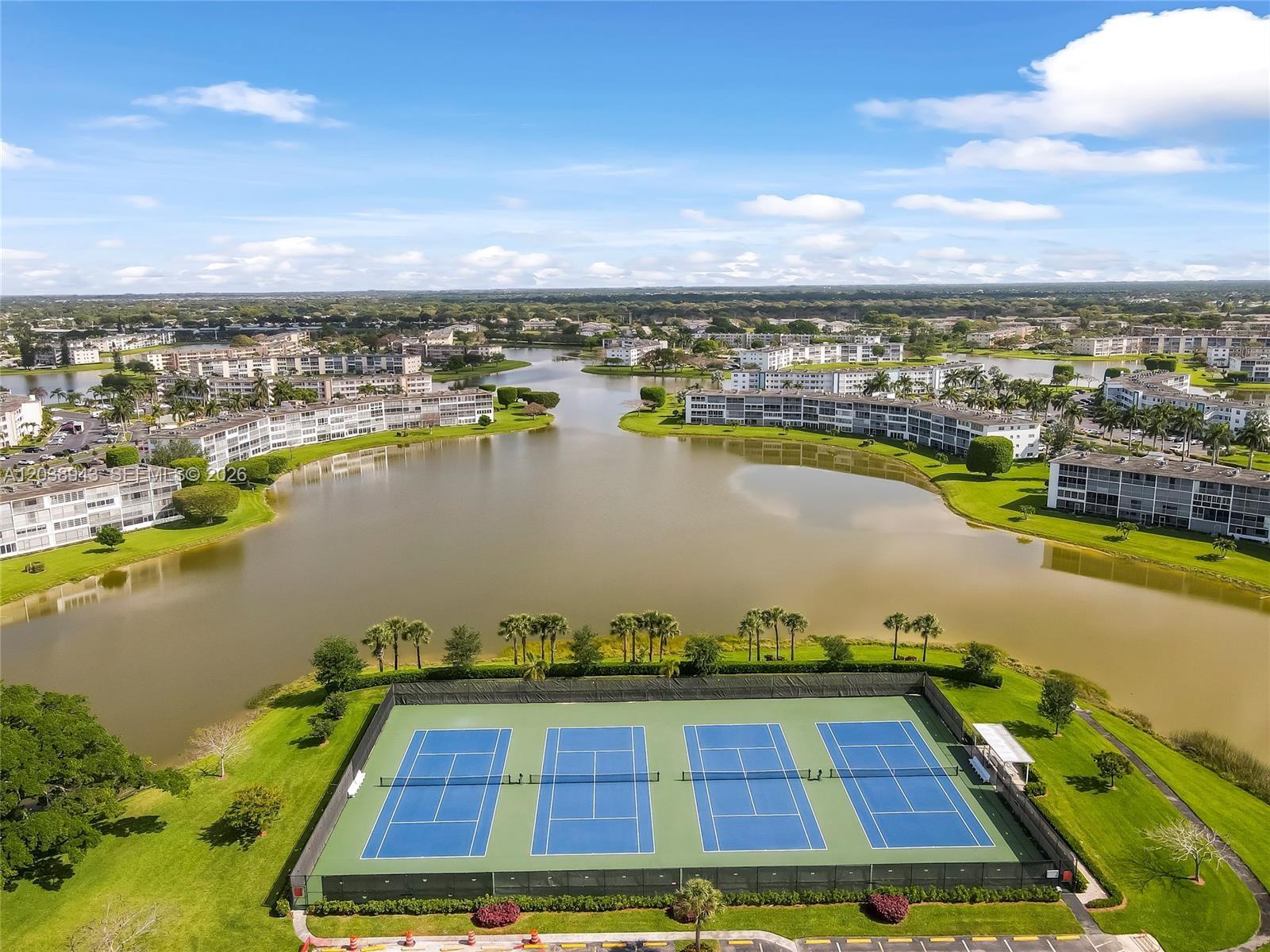 An aerial view shows four tennis courts situated next to a lake and apartment buildings — 82 Dorset B # B, Boca Raton FL 33434, BOCA RATON, FL