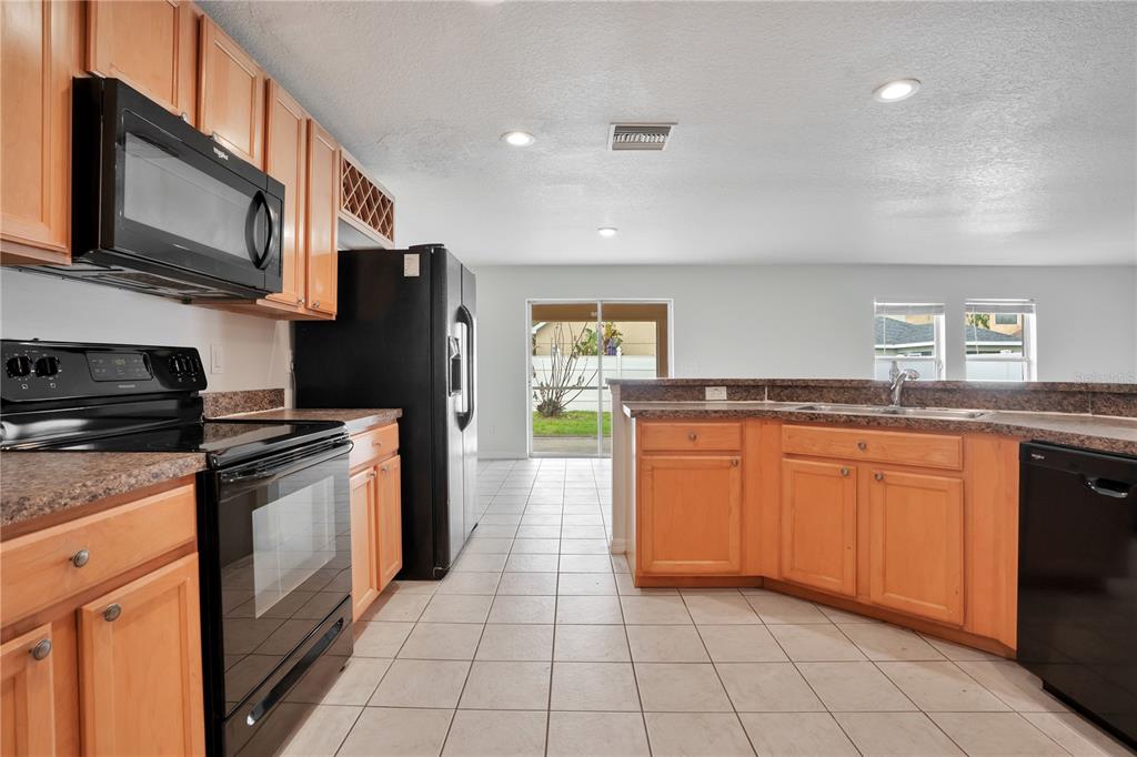 A kitchen with light wood cabinets, black appliances, and tile flooring leads to a sliding door — 12012 CITRUS LEAF DRIVE, GIBSONTON, FL