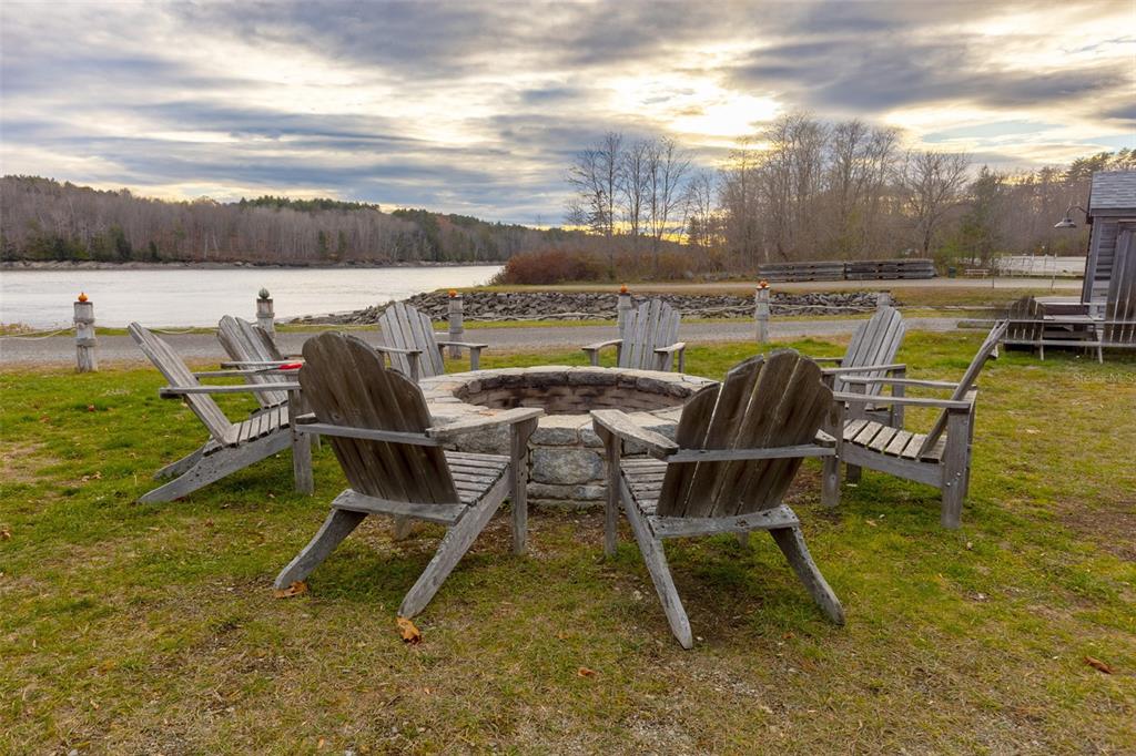 A stone fire pit surrounded by wooden Adirondack chairs overlooks a river at sunset — 108 MARINA ROAD, HAMPDEN, ME
