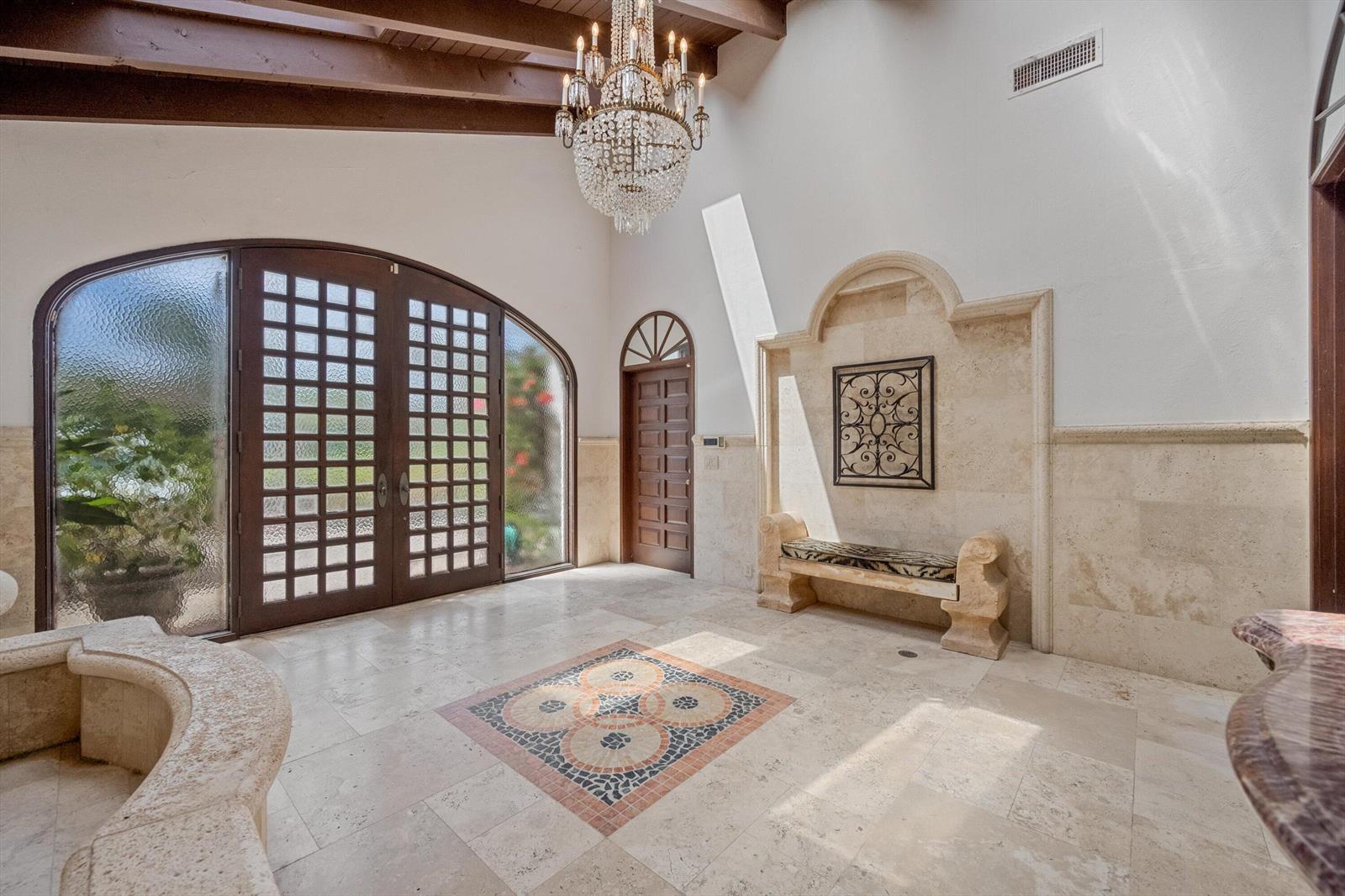 A grand entryway features a crystal chandelier, stone tile flooring, and a decorative stone bench — 23 Sloans Curve Drive, Palm Beach FL 33480, PALM BEACH, FL