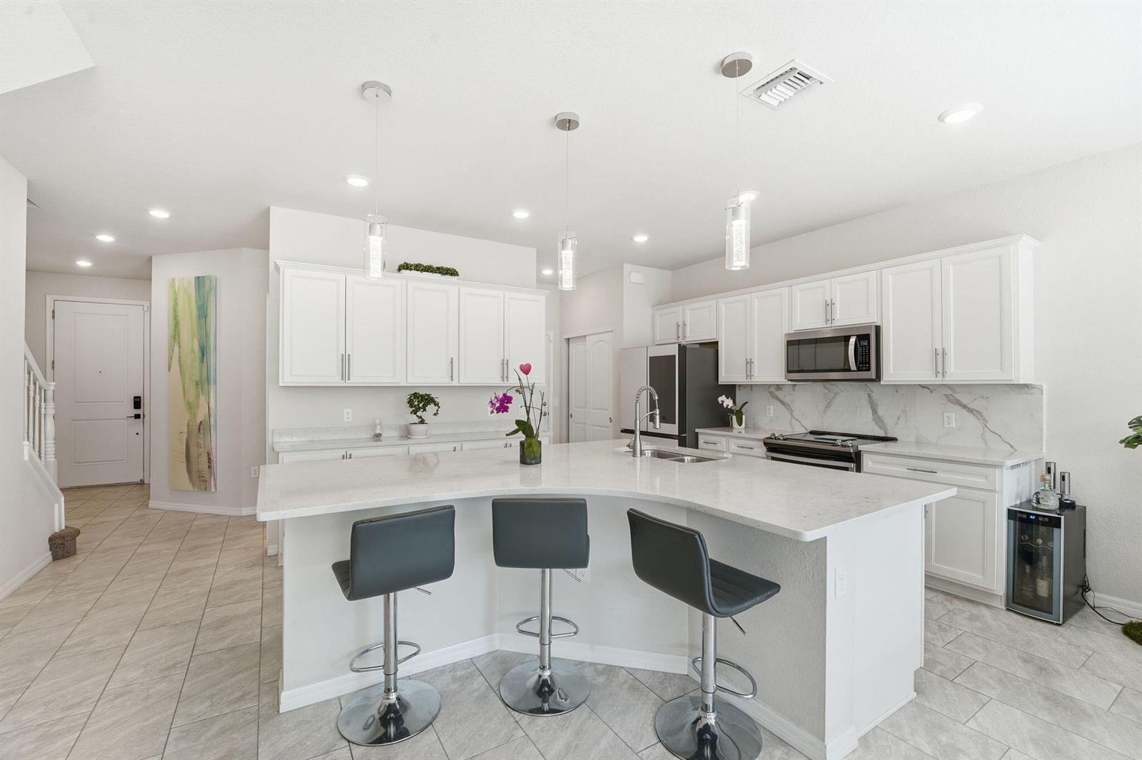 A modern kitchen features white cabinetry, a curved quartz island, and three gray bar stools — 4802 Blistering Way, Lake Worth FL 33467, LAKE WORTH, FL