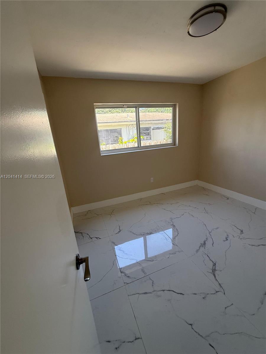 An empty room features white marble-patterned tile flooring, beige walls, and a single window — 2600 NW 112th St # 1, Miami FL 33167, MIAMI, FL