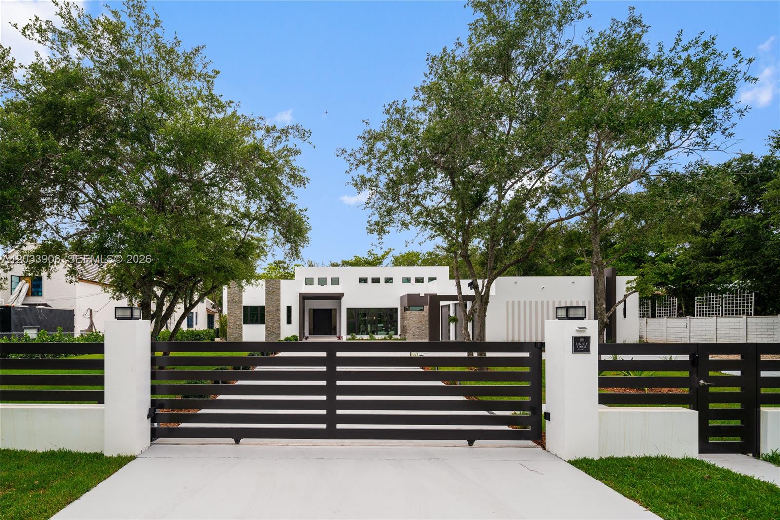 A modern white house with a flat roof is viewed through a black metal gate — 6200 SW 83 Av, Miami FL 33143, MIAMI, FL