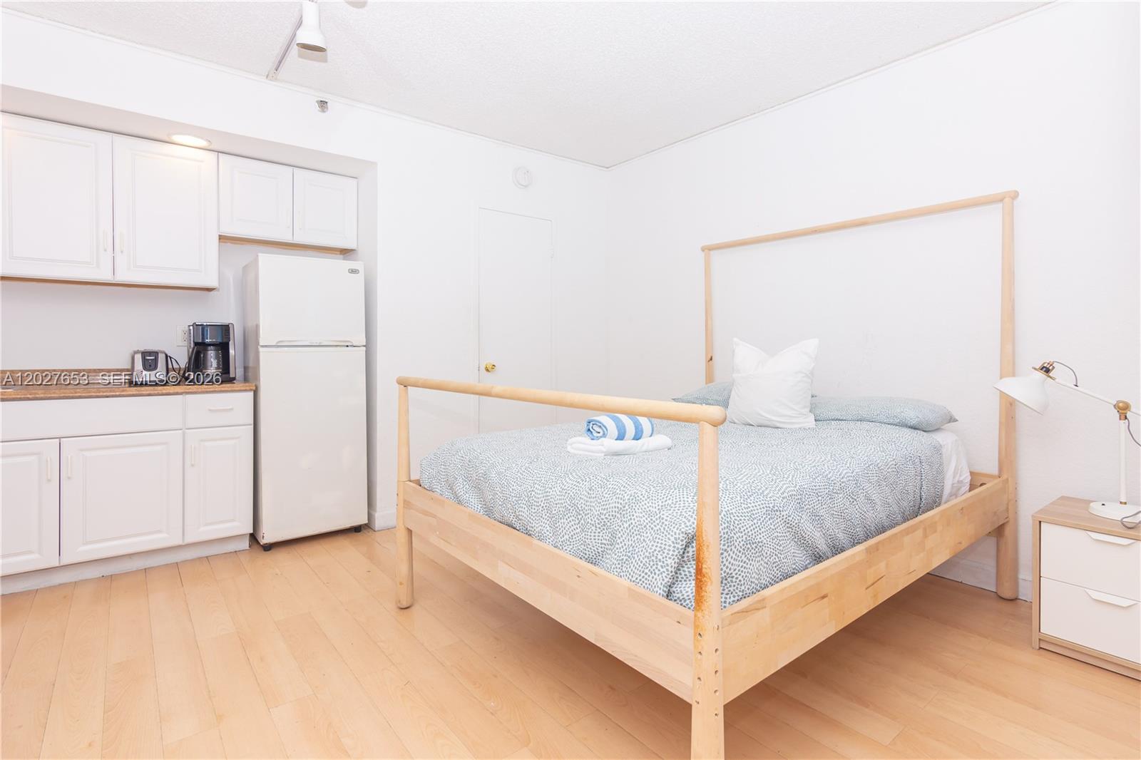 A studio apartment features a kitchenette with white cabinets, a refrigerator, and a wooden bed — 5445 Collins Ave # 1105, Miami Beach FL 33140, MIAMI BEACH, FL