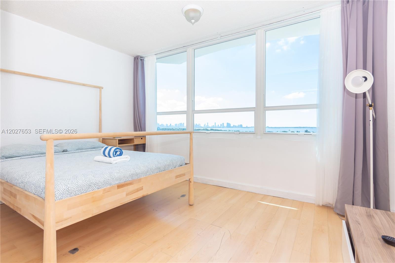 A bedroom features a light wood bed frame, hardwood floors, and large windows with water views — 5445 Collins Ave # 1105, Miami Beach FL 33140, MIAMI BEACH, FL
