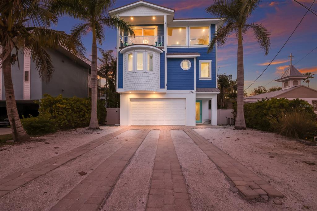 A three-story blue house with a white garage and palm trees at dusk — 206 CHURCH AVENUE, BRADENTON BEACH, FL