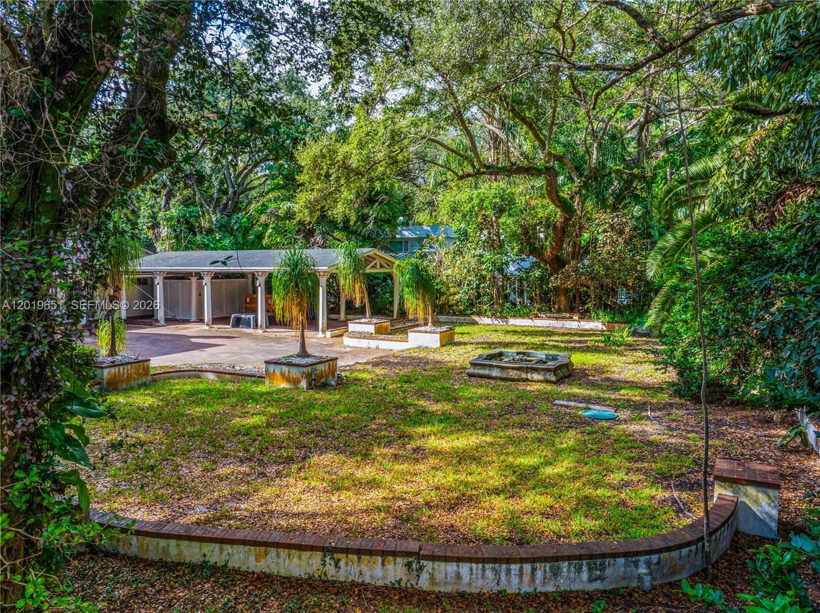 A backyard features a covered patio, a small fountain, and brick-edged planters under large trees — 3720 Poinciana Ave, Miami FL 33133, MIAMI, FL