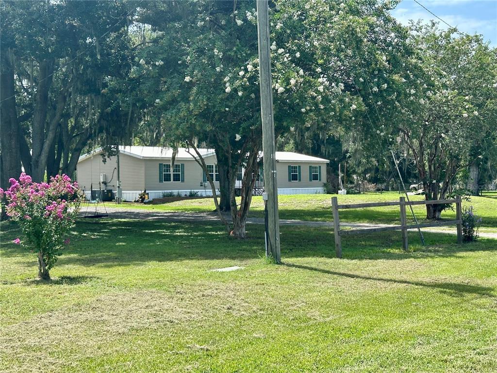 A single-story manufactured home sits on a grassy lot with trees and a wooden fence — 8528 AVON LADY PLACE, PLANT CITY, FL