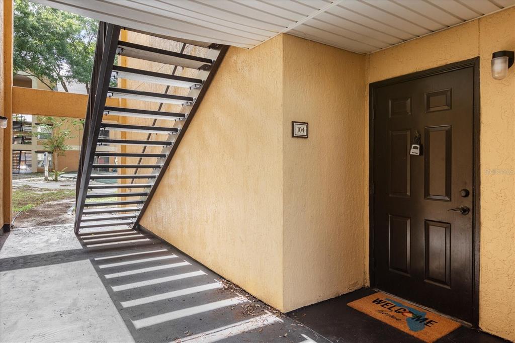 An exterior view of an apartment entrance with a staircase and a brown door — 7606 PISSARRO DRIVE UNIT 104, ORLANDO, FL