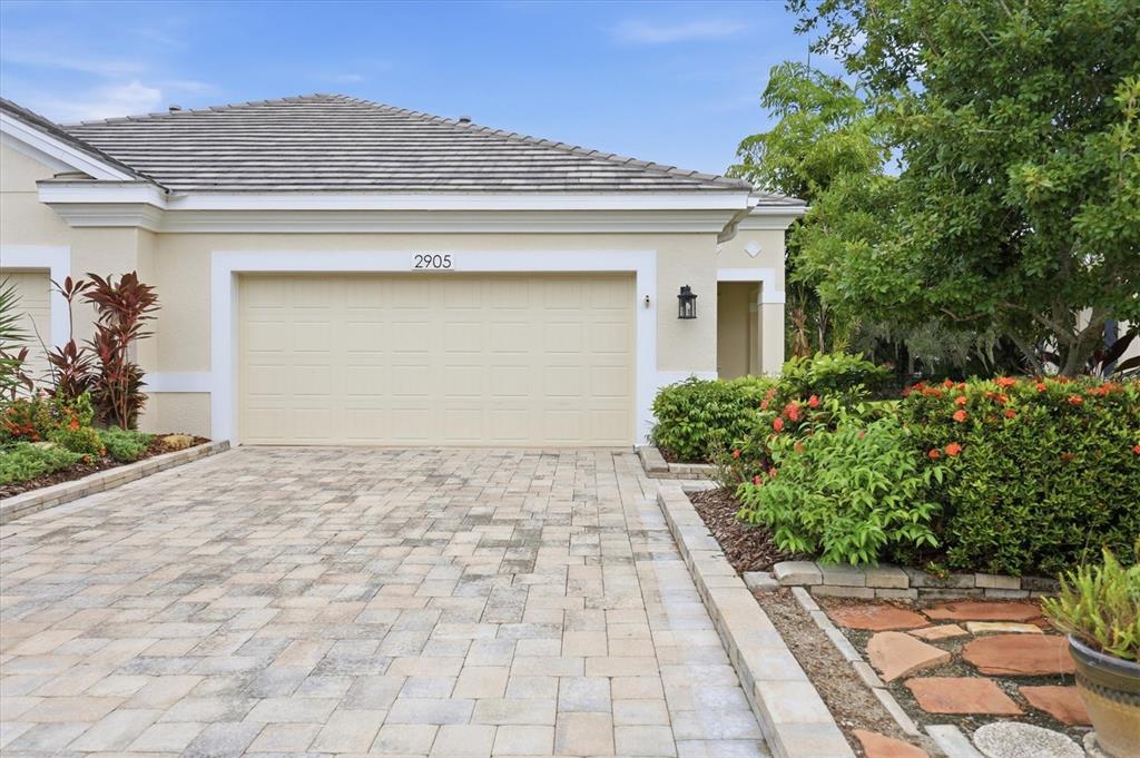 A beige house exterior features a two-car garage, a paved driveway, and landscaped garden beds — 2905 TRUSTEE AVENUE, SARASOTA, FL