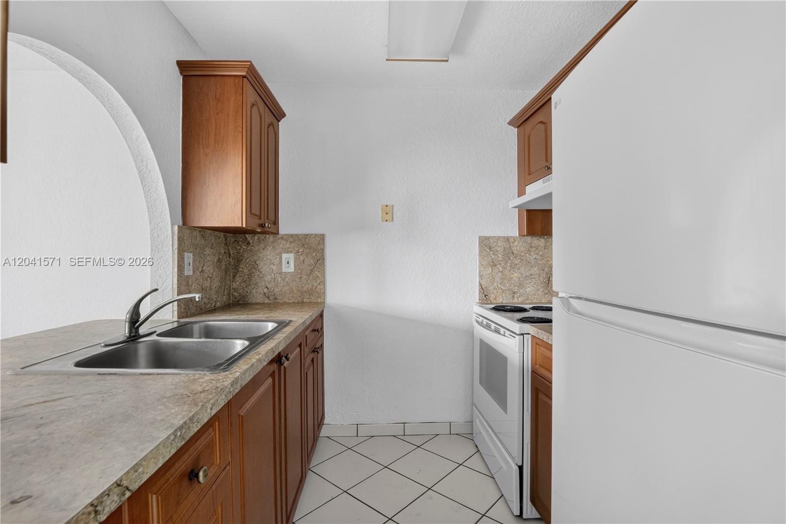 A galley kitchen features wood cabinets, stone countertops, white appliances, and white tile flooring — 5505 NW 7th St # W404, Miami FL 33126, MIAMI, FL