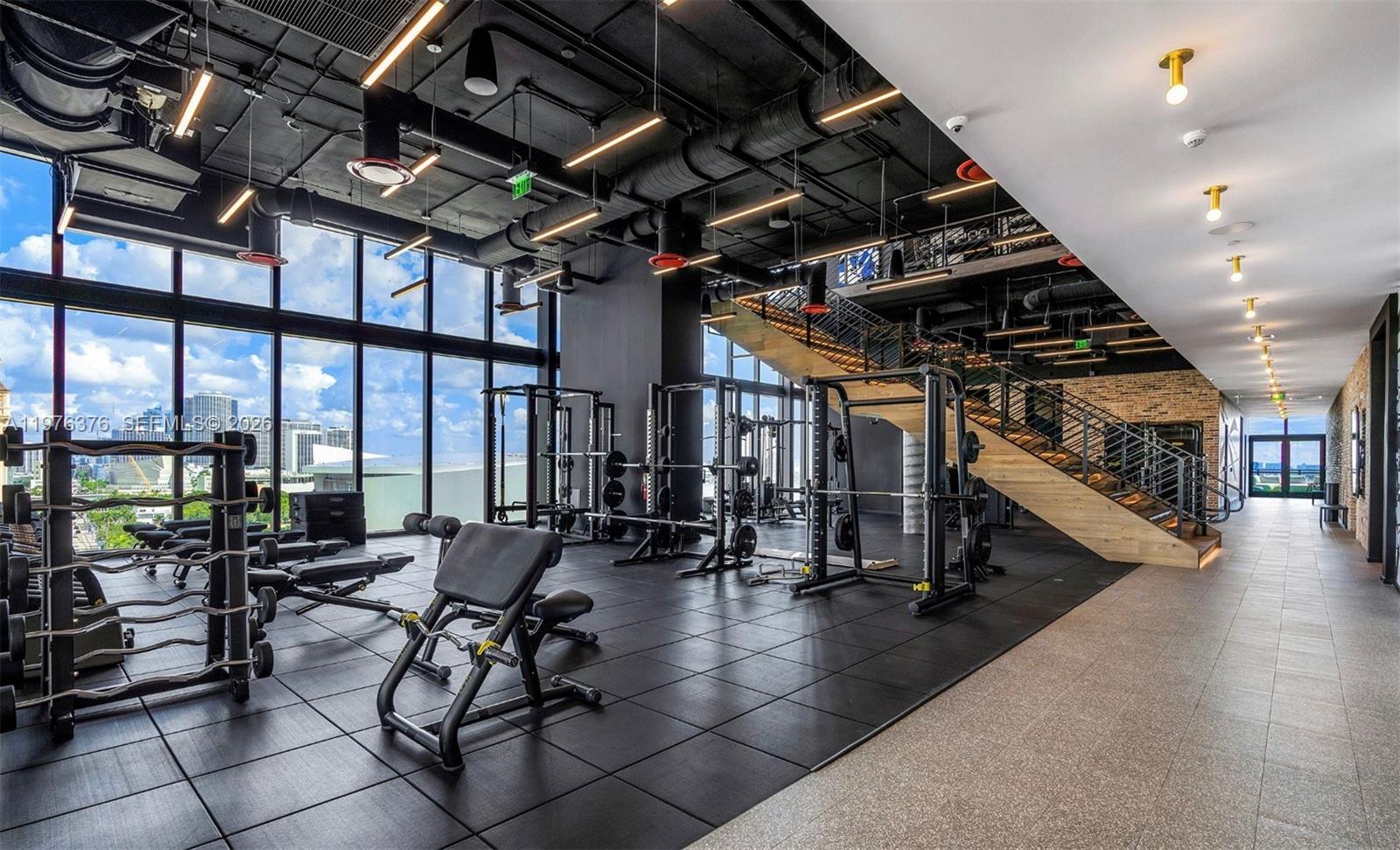 A modern gym features black rubber flooring, industrial ceilings, weight racks, and a wooden staircase — 398 NE 5th St # 4701, Miami FL 33132, MIAMI, FL