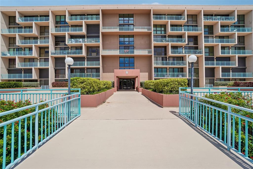 A concrete walkway leads to the entrance of a multi-story apartment building with blue railings — 1 KEY CAPRI UNIT 406W, ST PETERSBURG, FL