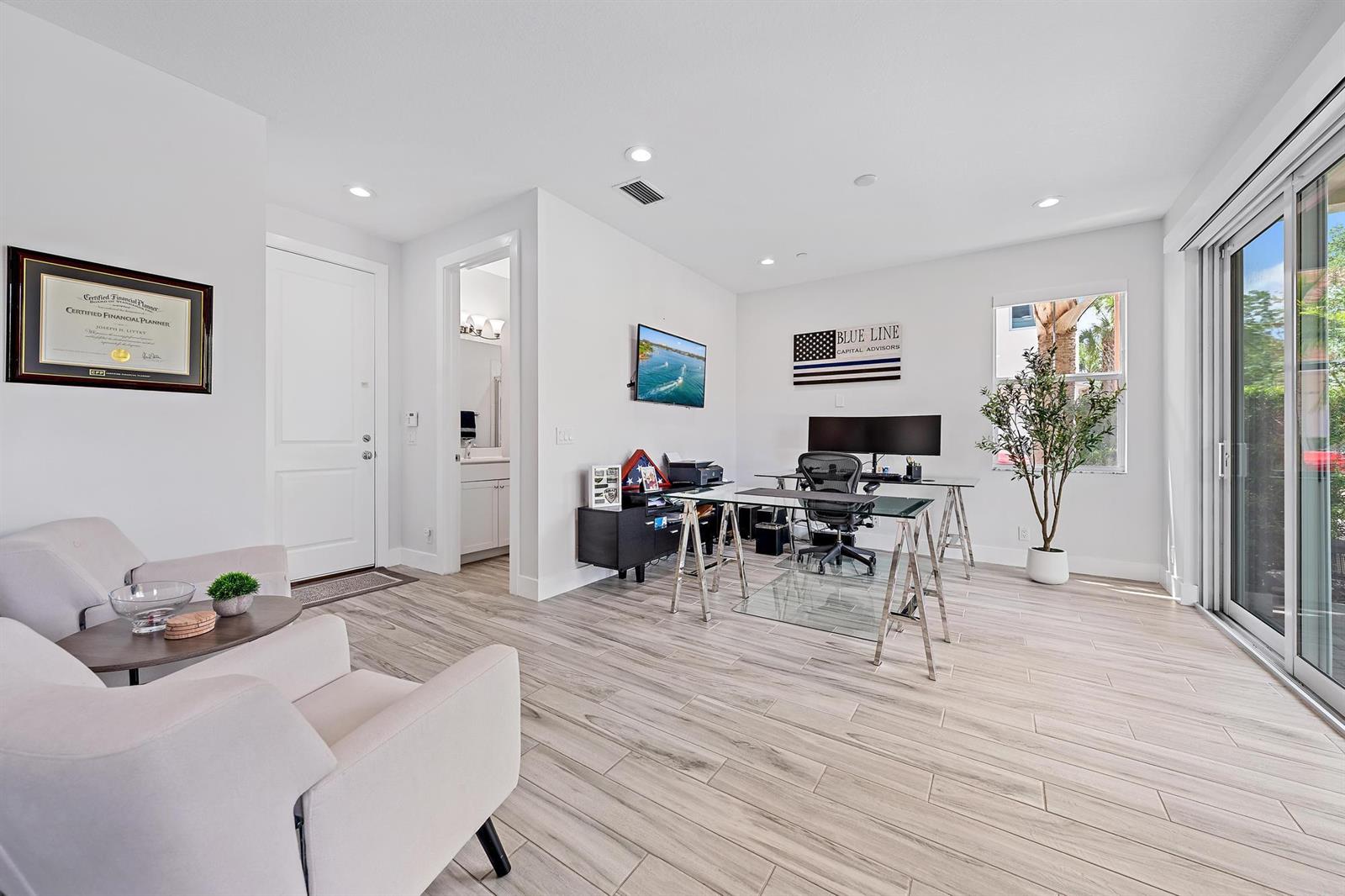 A bright home office features light wood-look tile flooring, a glass desk, and sliding glass doors — 4145 Faraday Way, Palm Beach Gardens FL 33418, PALM BEACH GARDENS, FL