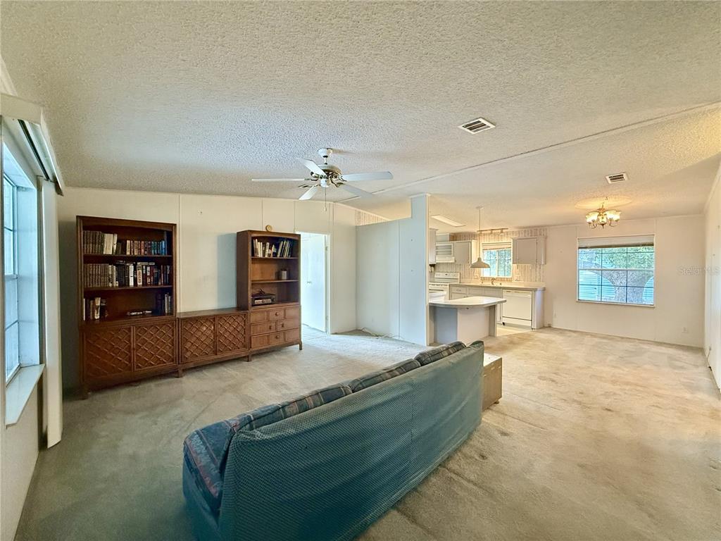 A living room with carpeted floors, a blue sofa, and wooden shelving units leads to a kitchen — 5152 FOREST GLENN DRIVE, SPRING HILL, FL