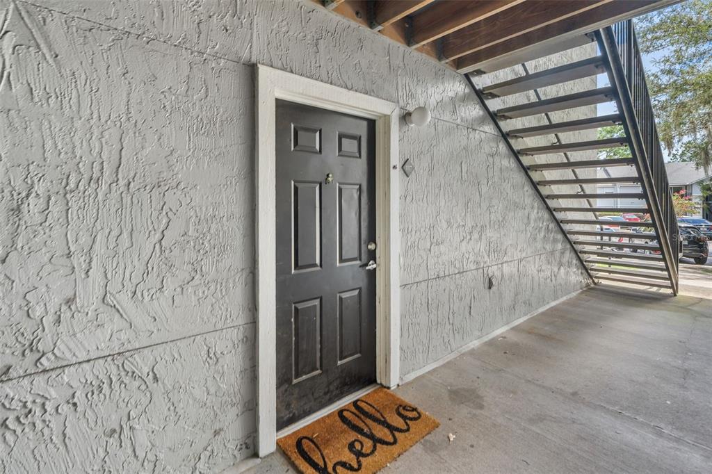 A dark front door with a welcome mat sits beneath an outdoor staircase — 3705 SW 27TH STREET UNIT 1117, GAINESVILLE, FL