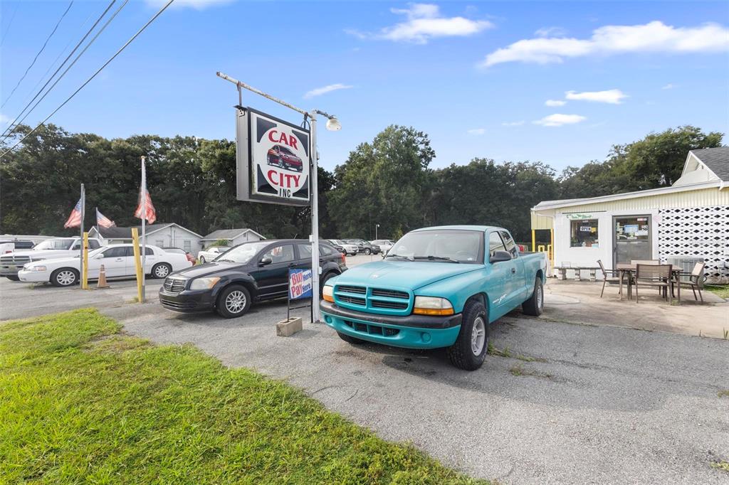 A teal pickup truck is parked in front of a car dealership with a sign — 2490 N WOODLAND BOULEVARD, DELAND, FL