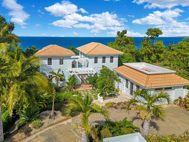 An aerial view shows a light blue two-story house with a detached garage near the ocean — 1 COLINA LINDA- "TRANQUILO", RINCON, PR