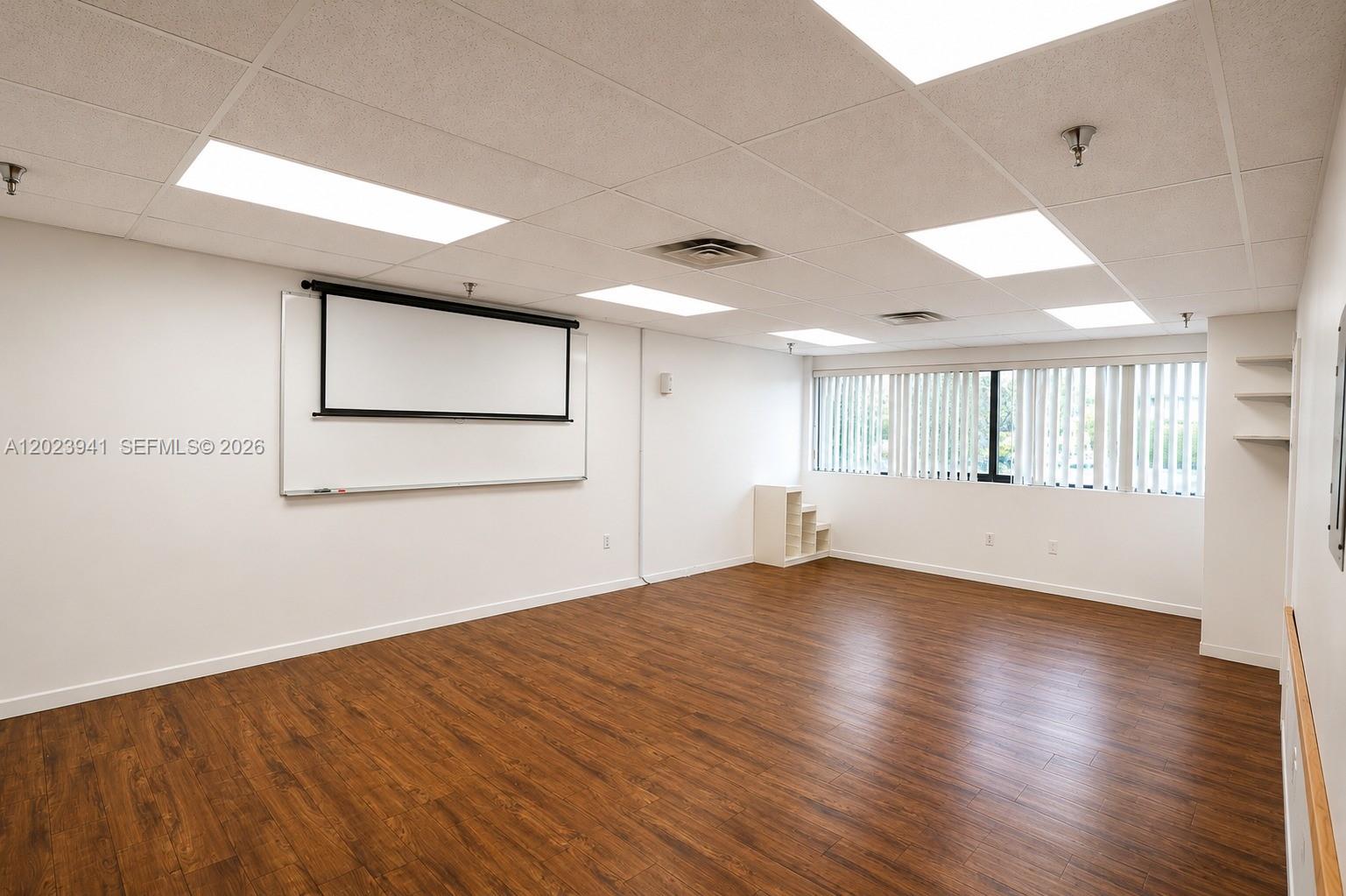 An empty office room features wood-look flooring, a projector screen, a whiteboard, and large windows — 4355 W 16th Ave # 211, Hialeah FL 33012, HIALEAH, FL