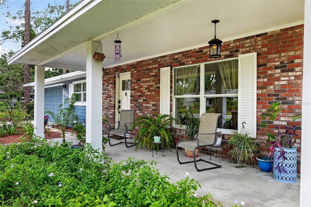 A brick house exterior features a covered front porch with two chairs and potted plants — 3862 NW 36TH PLACE, GAINESVILLE, FL