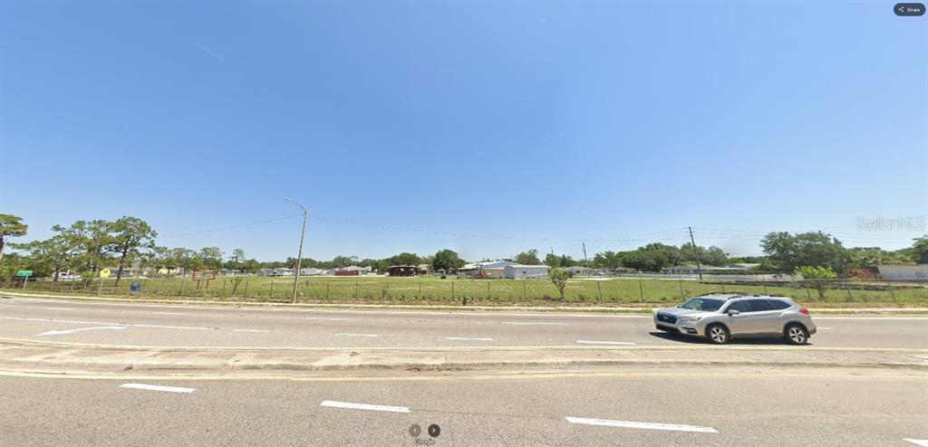 A silver SUV drives past a large, fenced, vacant grassy lot under a clear blue sky — HWY 60 E, LAKE WALES, FL