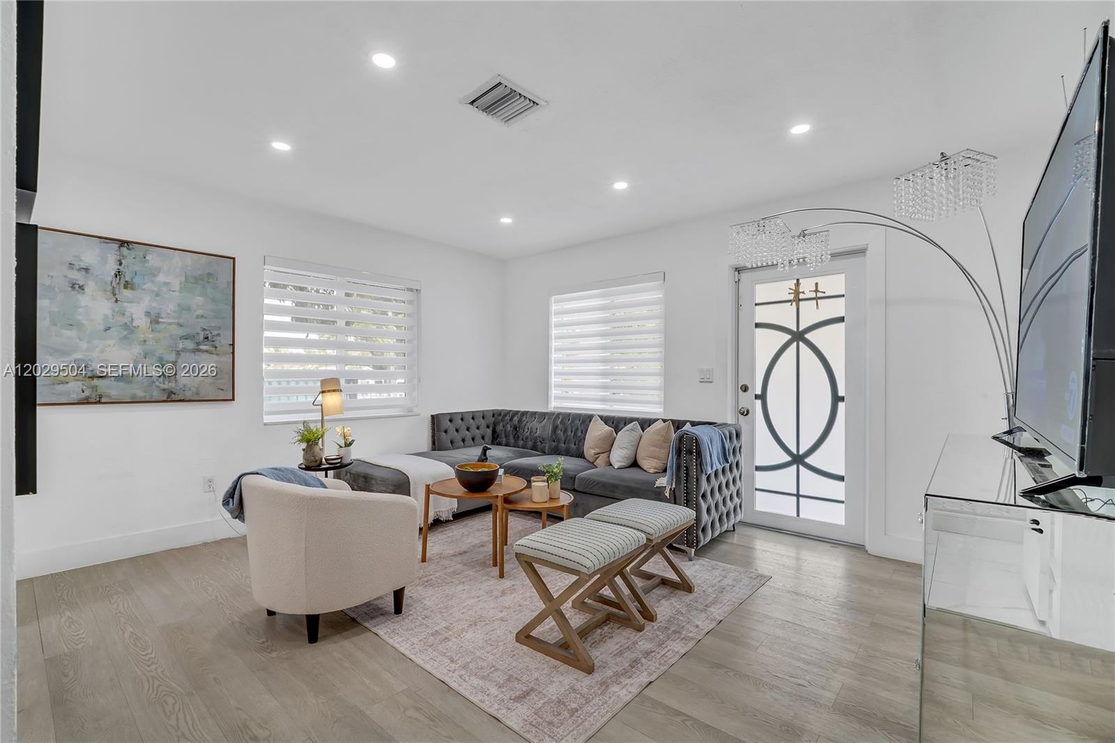 A modern living room features a grey tufted sectional, a white armchair, and light wood flooring — 6404 Meade St, Hollywood FL 33024, HOLLYWOOD, FL