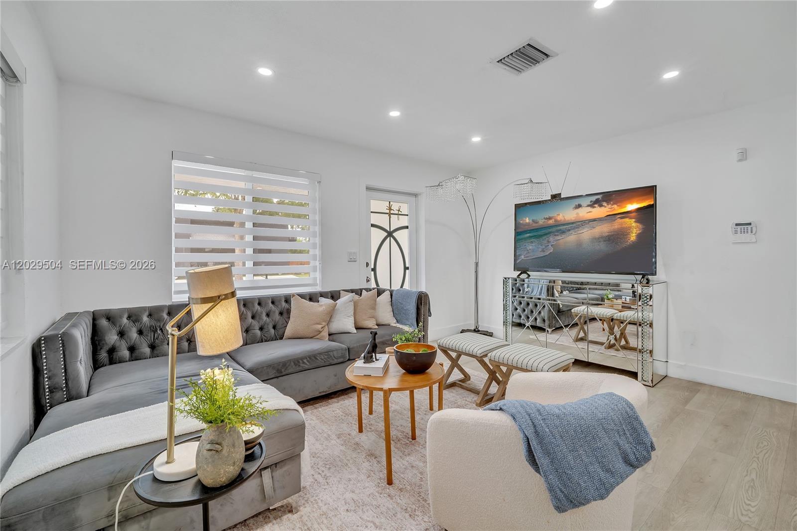 A living room features a grey tufted sectional sofa, a mirrored TV stand, and white walls — 6404 Meade St, Hollywood FL 33024, HOLLYWOOD, FL