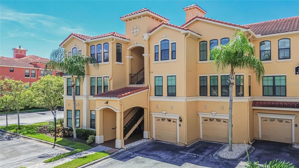 A multi-story tan stucco apartment building features a tiled roof, arched entryways, and attached garages — 2723 VIA CAPRI UNIT 832, CLEARWATER, FL