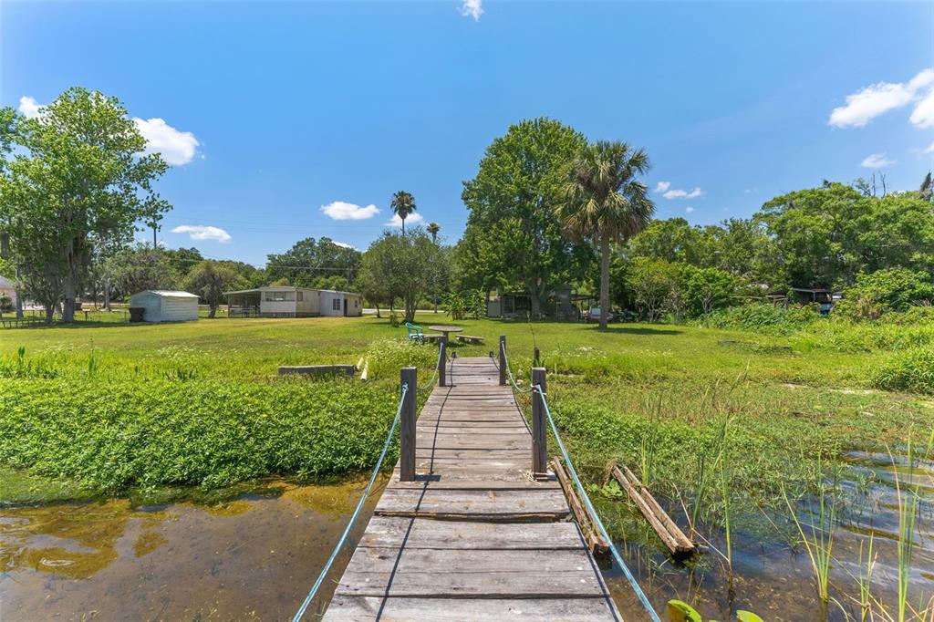 A wooden dock extends from the water toward a grassy yard with trees and buildings — 7570 BERKLEY ROAD, POLK CITY, FL