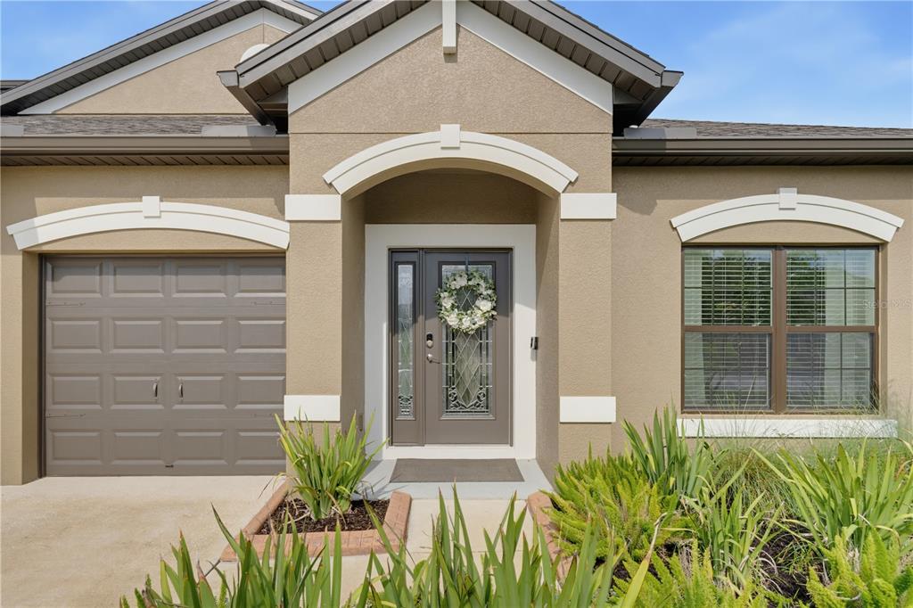 The exterior of a tan house features a garage, a front door with a wreath, and landscaping — 7903 LAGO MIST WAY, WESLEY CHAPEL, FL