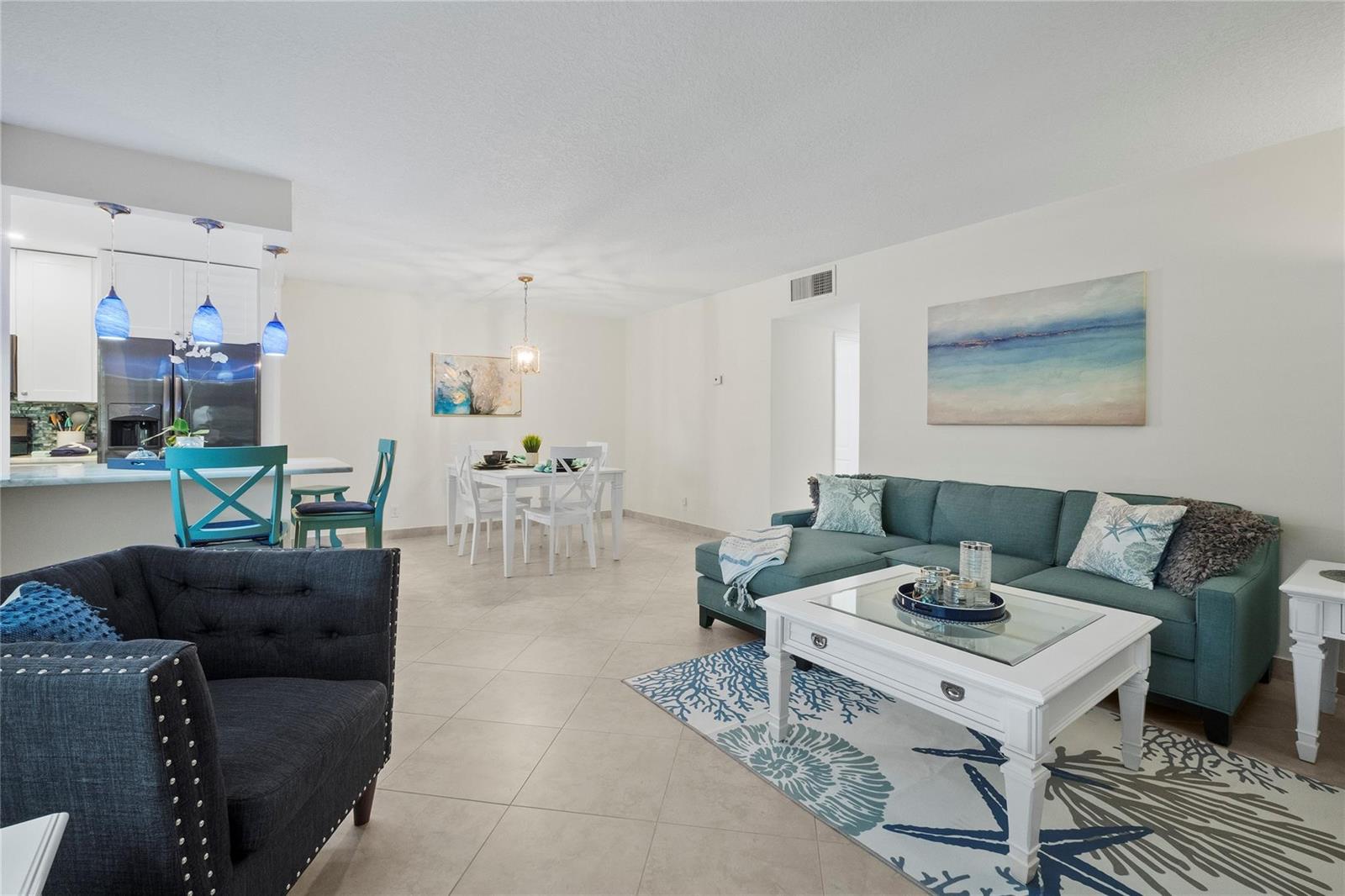 An open-concept living and dining area features tile flooring, a teal sectional, and white furniture — 1340 NE 28th Avenue # 242, Pompano Beach FL 33062, POMPANO BEACH, FL