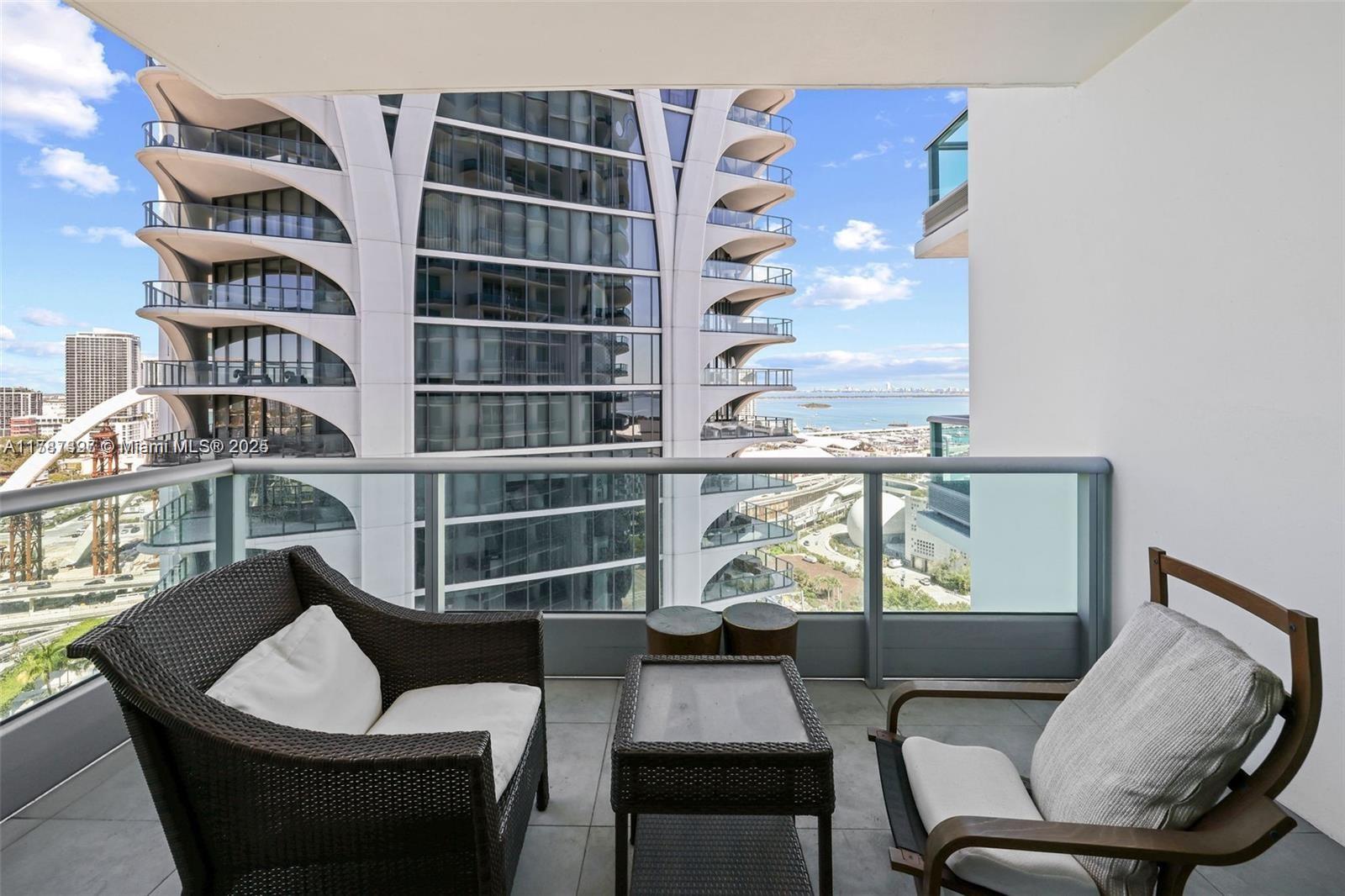 A balcony with wicker furniture overlooks a modern high-rise building and a distant bay — 900 Biscayne Blvd # 2410, Miami FL 33132, MIAMI, FL