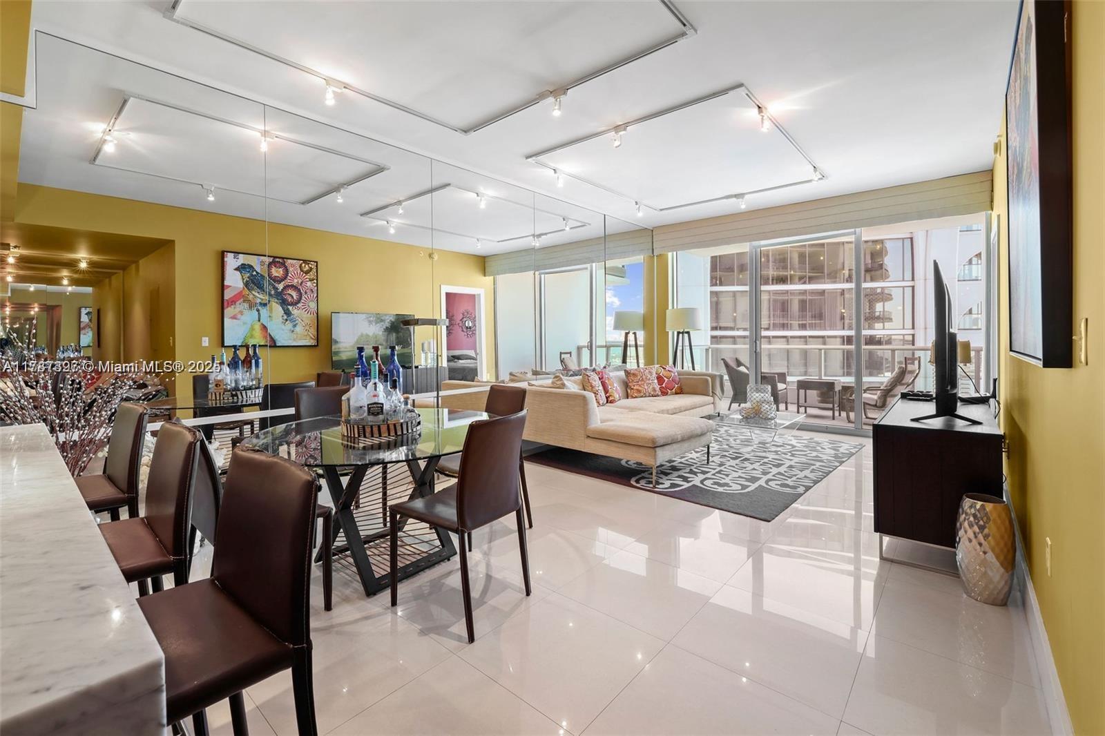A modern living and dining area features yellow walls, mirrored accents, tile flooring, and a balcony — 900 Biscayne Blvd # 2410, Miami FL 33132, MIAMI, FL
