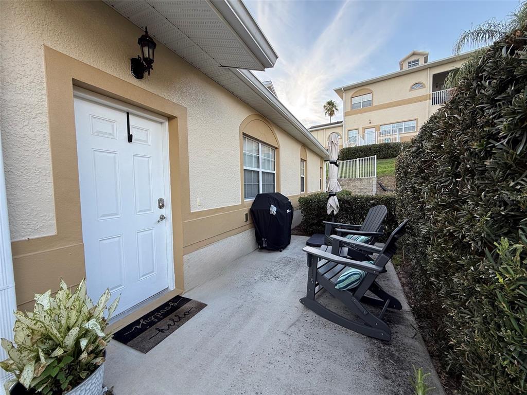 A white door opens onto a concrete patio with two rocking chairs and a grill — 17411 PROMENADE DRIVE, CLERMONT, FL
