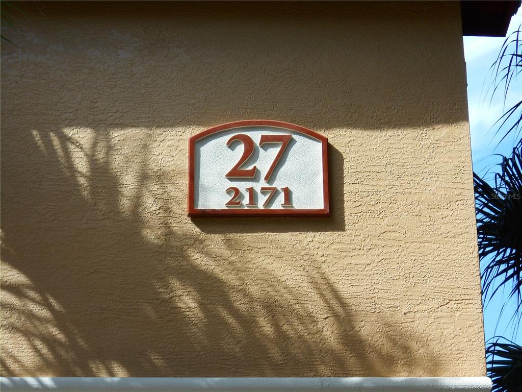 A building exterior wall features a plaque displaying the numbers 27 and 2171 — 2171 PORTOFINO PLACE UNIT 2727, PALM HARBOR, FL