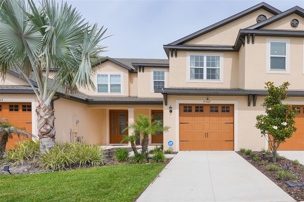 A two-story townhouse exterior with tan stucco walls, orange garage doors, and palm trees — 11367 CAY SPRUCE WAY, SAN ANTONIO, FL