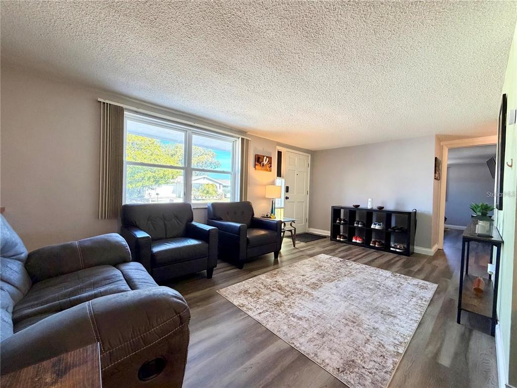 A living room features gray wood-look flooring, a textured ceiling, and dark leather seating — 3118 ELKRIDGE DRIVE, HOLIDAY, FL