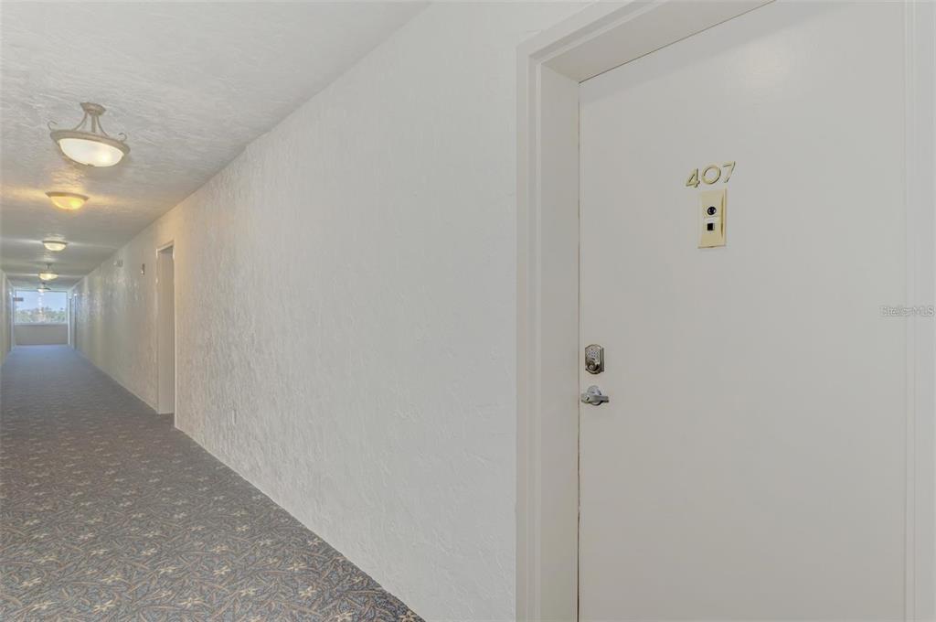 A long apartment hallway features white walls, patterned carpet, and a door labeled 407 — 6114 43RD STREET W UNIT 407E, BRADENTON, FL