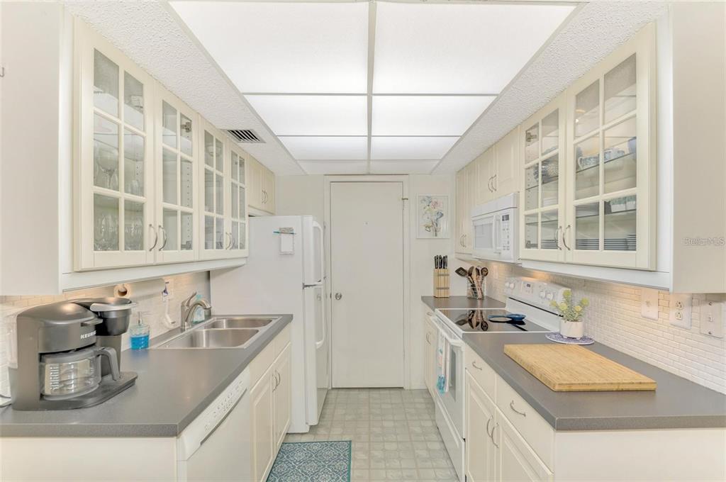 A galley kitchen features white cabinets with glass-front doors, gray countertops, and a white refrigerator — 6114 43RD STREET W UNIT 407E, BRADENTON, FL