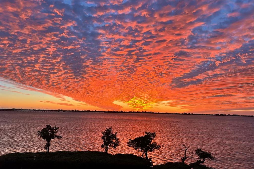A vibrant sunset sky glows orange and red over a calm body of water with trees — 264 SAINT LUCIA DRIVE UNIT 201, BRADENTON, FL